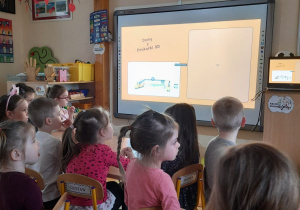 Dzieci siedzą na dywanie tyłem oglądają prezentację multimedialną
