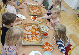 dzieci siedzą przy stolikach układają warzywa wędlinę i ser na cieście od pizzy