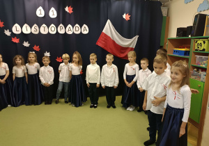dzieci ubrane na galowo stoją w półkolu na tle napisu 11 listopada oraz flagi biało-czerwonej