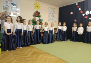 dzieci ubrane na galowo stoją w półkolu na tle napisu 11 listopada oraz flagi biało-czerwonej
