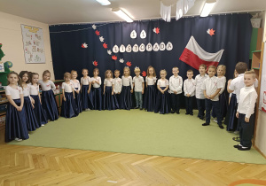 dzieci ubrane na galowo stoją w półkolu na tle napisu 11 listopada oraz flagi biało-czerwonej