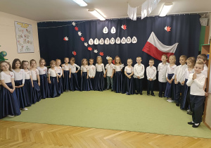 dzieci ubrane na galowo stoją w półkolu na tle napisu 11 listopada oraz flagi biało-czerwonej