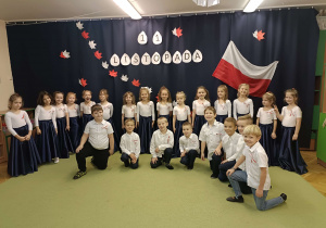dzieci ubrane na galowo stoją w półkolu na tle napisu 11 listopada oraz flagi biało-czerwonej