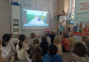 dzieci siedzą przed tablicą multimedialną na której jest samochód na przejeździe kolejowym